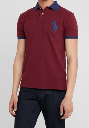 Homme portant un polo bordeaux avec col et poignets bleu marine, logo joueur de polo bleu sur la poitrine et jean bleu foncé.