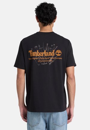 Mand iført sort Timberland t-shirt med orange logo og værktøjstegninger på ryggen, kombineret med lyseblå jeans, med ryggen mod.