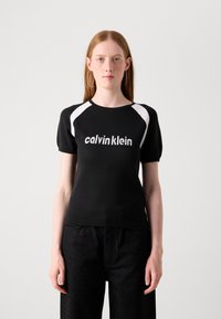 Juodas megztinis su baltais raglano rankovėmis, akcentuojantis ryškų "calvin klein" logotipą. Prigludusi išvaizda; trumpomis rankovėmis; lygus paviršius.