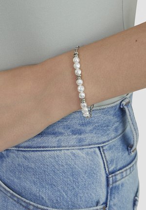 Armband mit abwechselnden weißen Perlen und silbernen Kugeln am Handgelenk, kombiniert mit hellblauen Jeans und grauem Oberteil.