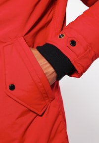 Veste isolée rouge avec une finition brillante, dotée d'un poignet à côtes noir et de détails de fermeture à pression sur les manches et la poche.