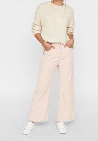 Modell i en krämfärgad tröja och ljusrosa vida byxor står med händerna på höfterna, ihop med vita sneakers mot en neutral bakgrund.