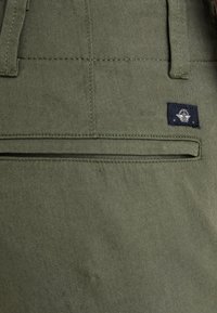 Shorts en tissu vert olive avec une poche arrière zippée et une petite étiquette de marque. Couture robuste avec une texture lisse.