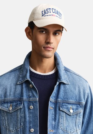 Jonge man met een beige pet met de tekst "EAST COAST", een marineblauwe trui en een lichtblauwe spijkerjas, die naar voren kijkt met een neutrale uitdrukking.