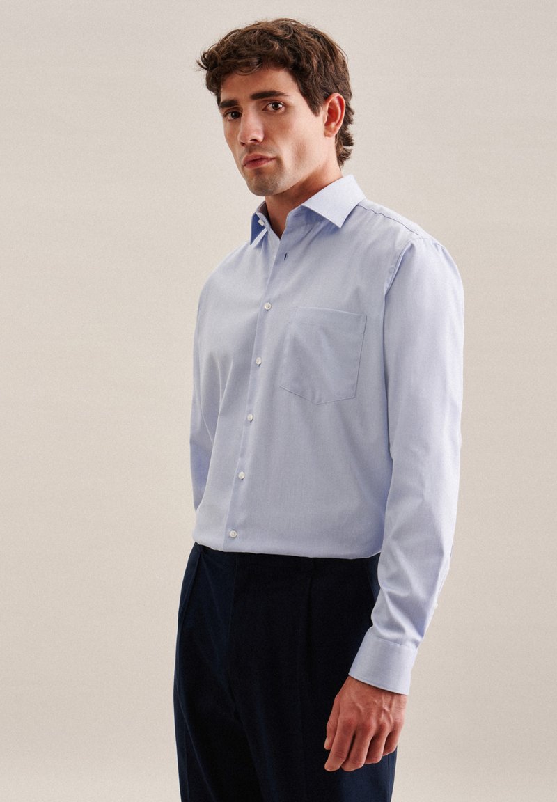 Chemise bleu clair à col pointu, avec une poche poitrine unique, des manches longues et des boutons blancs, portée avec un pantalon sombre.