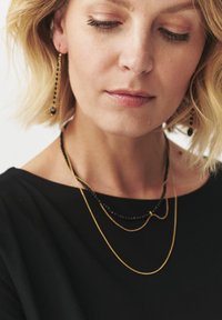 Gold and black layered necklaces with beaded accents worn with a black top. Simple earrings complement the jewelry. Smooth textures visible.