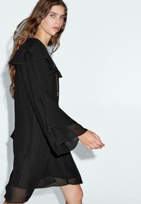 Schwarzes Kleid mit einem rüschenden Ausschnitt und fließenden, durchsichtigen Ärmeln. Der Stoff ist leicht und hat eine glatte Textur, mit knielangem Schnitt.