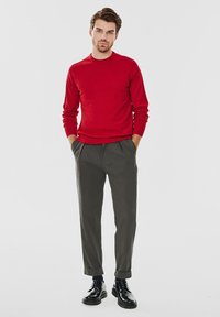 Czerwony sweter z okrągłym dekoltem zestawiony z ciemnoszarymi, zwężanymi spodniami. Model nosi czarne buty sznurowane o błyszczącej powierzchni.