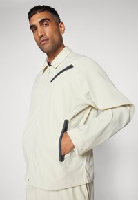 Light beige zip-up jacket with a textured fabric, featuring a black pocket accent and side pockets. The design includes a collar and long sleeves.
