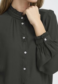 Dunkelgrün Bluse mit hohem Rüschkragen, langen Ärmeln und elastischen Bündchen. Verfügt über eine durchgehende Knopfleiste mit großen weißen Knöpfen.