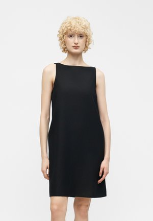 Femme blonde aux cheveux bouclés portant une robe noire sans manches jusqu'aux genoux sur un fond clair uni.