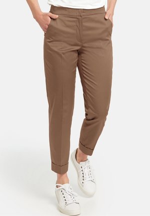 Femme portant un pantalon slim marron avec des ourlets retournés et des baskets blanches, mains dans les poches, debout sur un fond blanc.