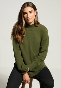 Grüner gestrickter Pullover mit hohem Kragen, gerippten Bündchen und Saum. Zum schwarzen Jeans getragen, Modell sitzt auf einem Holzstuhl.