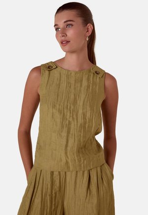 Femme aux cheveux bruns attachés en queue de cheval, portant un haut sans manches texturé vert olive avec des détails de boutons sur les épaules et un pantalon plissé assorti.