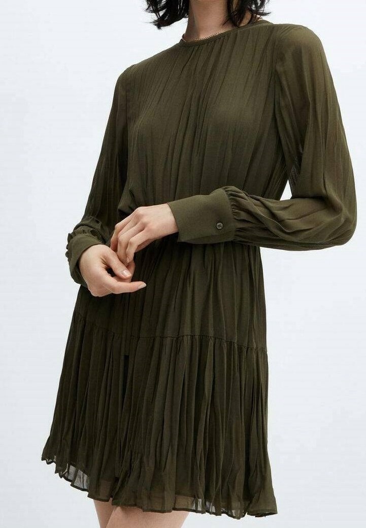 Femme portant une robe plissée verte olive à manches longues avec des poignets boutonnés et un col rond, debout devant un fond uni.