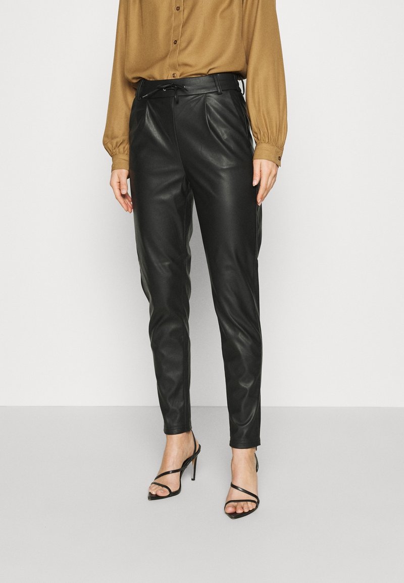 Svarta läderbyxor med en elegant design, featuring en bältad midja, raka ben och sidofickor. Kombination med en brun blus och svarta klackar.