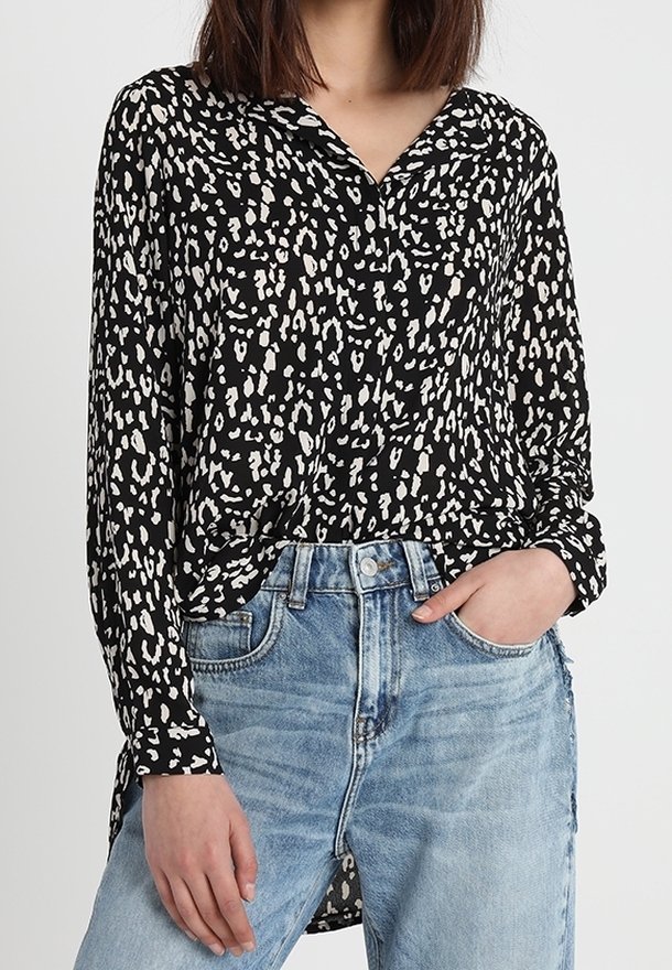 Schwarze und weiße gemusterte Bluse mit Kragen, Knopfleiste und langen Ärmeln, kombiniert mit hellblauen, hoch geschnittenen Jeans.