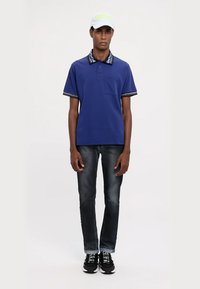 Modrá polo košeľa s vzorovaným golierom, krátkymi rukávmi a vreckom na hrudi, skombinovaná s tmavými džínsami a čiernymi teniskami. Biela čiapka s logom.