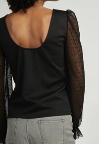 Top nero con scollo profondo sul retro, corpino aderente e maniche a palloncino sheer nere con pois a rilievo. Abbinato a pantaloni grigi.