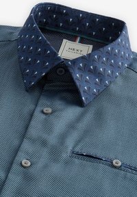 Cămașă albastră cu un model texturat, având un guler bleumarin cu un design geometric și detalii cu nasturi la față și la buzunar.