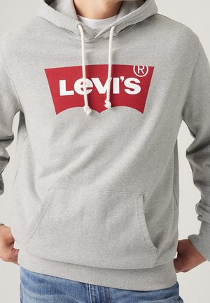 Mężczyzna w jasnoszarej bluzie z kapturem z białymi sznurkami i czerwonym logo Levi's na klatce piersiowej, w połączeniu z niebieskimi dżinsami.