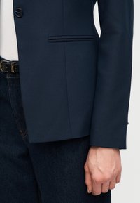 Gros plan sur une personne portant un blazer bleu marine, un haut blanc, un jean foncé avec une ceinture noire, et la main détendue le long du corps.