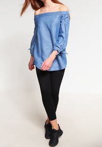 Kvinna som bär en blå off-shoulder-blus med knutna ärmar, svarta leggings och svarta sneakers, stående mot en enkel ljus bakgrund.