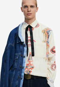 Camicia bianca con motivi colorati, cravatta nera e taschino; sovrapposta a una giacca di denim blu. Il denim ha grandi bottoni e un colletto blu chiaro.