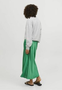 Felpa grigia, gonna maxi in satin verde, vestibilità casual con un design fluido. Sandali neri. Capelli ricci visibili da dietro, sfondo neutro.