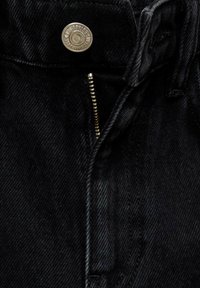 Jean en denim noir avec une fermeture éclair dorée partiellement ouverte et un bouton argenté gravé d’un texte sur la ceinture.