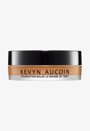 Kevyn Aucoin Foundation Balm in einem runden Glas, mit einem mattschwarzen Deckel, einem transparenten Boden und einem cremigen beigefarbenen Produkt, das sichtbar darin ist.