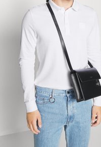 Vit långärmad pikétröja med krage, tillsammans med ljusblå jeans och en svart läderaxelväska.