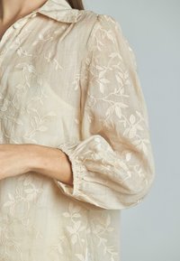 Blusa leggera color crema con ricami floreali, colletto e polsini elasticizzati. Tessuto semi-trasparente con trame sottili.