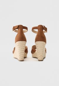 Sandali con tacco wedge in suede marrone, con tacco in juta intrecciata, cinturino alla caviglia con fibbia e design aperto sulla punta. Il tacco texturizzato aggiunge dettagli.