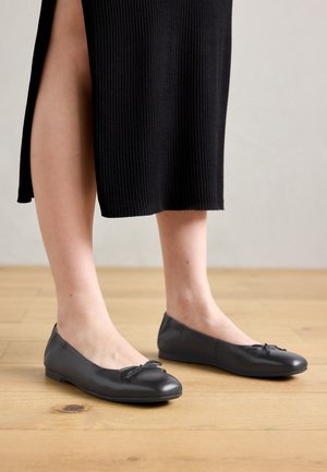Bare legs wearing black leather ballet flats with small bows, paired with a black ribbed skirt with a side slit, standing on wooden floor.