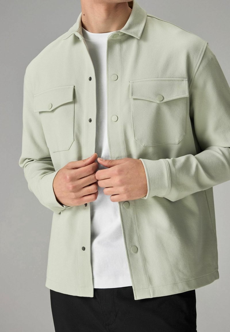 Homme portant une veste boutonnée vert clair sur une chemise blanche, en train de boutonner les boutons, debout devant un fond gris uni.