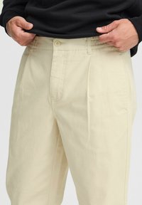 Beige geplooide broek van katoen, met een knoopsluiting, zijzakken en een ontspannen pasvorm. De stof heeft een gladde textuur.
