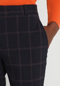 Henkilö, joka on pukeutunut tummansinisiin ruutuhousuihin ja oranssiin pitkähihaiseen paita, käsi housujen taskussa.