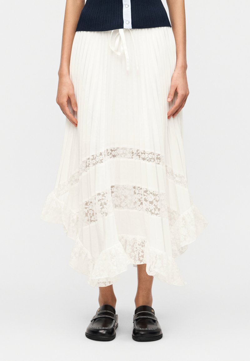 Γυναίκα που φοράει λευκή πλισέ midi φούστα με δαντ ελένια πάνελ και βολάν στο τελείωμα, συνδυασμένη με μαύρα μοκασίνια και μπλε ναυτικό αμάνικο πουλόβερ.