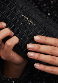 Schwarze vegane Leder-Clutch mit einem Krokodilmuster und goldenen Markenlogo "ISABEL BERNARD PARIS." Die Hände, die die Clutch halten, sind sichtbar.