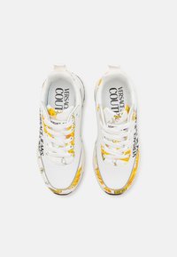 Hvite joggesko med blomstrete gult design, logo tekst på sidene, teksturerte snørebånd og polstrede såler, med en glatt overmateriale.