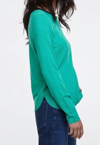 Femme portant un pull à manches longues vert vif avec des fentes sur les côtés et un jean bleu foncé, vue de côté sur un fond uni.
