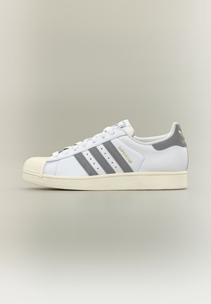 Baskets Adidas Superstar blanches avec des rayures grises, texte doré « Superstar » et semelle écrue sur un fond gris uni.