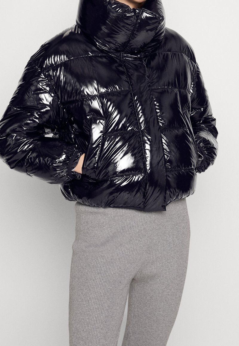 Personne portant une doudoune noire brillante avec un col haut et des poches, associée à un pantalon en maille côtelée gris clair.