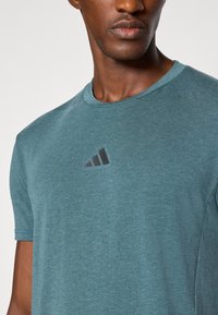 Camiseta de algodón verde-azul con cuello redondo y mangas cortas, con un logo negro de Adidas en el pecho.