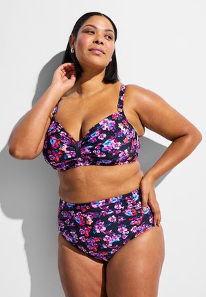 Žena v čiernych dvojdielnych plavkách s ružovými a modrými kvetinovými vzormi stojaca na bielom pozadí s jednou rukou na boku.