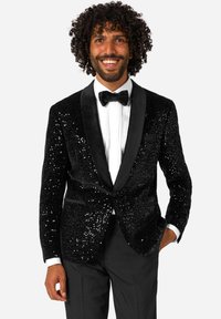 Moški v črnem bleščečem fraku, beli srajci, črni metuljčku in črnih hlačah, se smeji s poudarjeno roko v žepu.