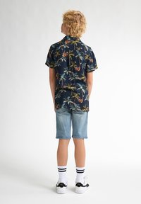 Cămașă cu mânecă scurtă, albastră închis, cu un model colorat de palmieri, asociată cu pantaloni scurți din denim de culoare bleu deschis și adidași negri cu șosete albe cu dungi.