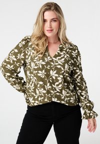Blouse vert olive avec motif floral blanc, dotée d'un décolleté en V, tissu texturé et poignets froncés. Associée à un pantalon noir.