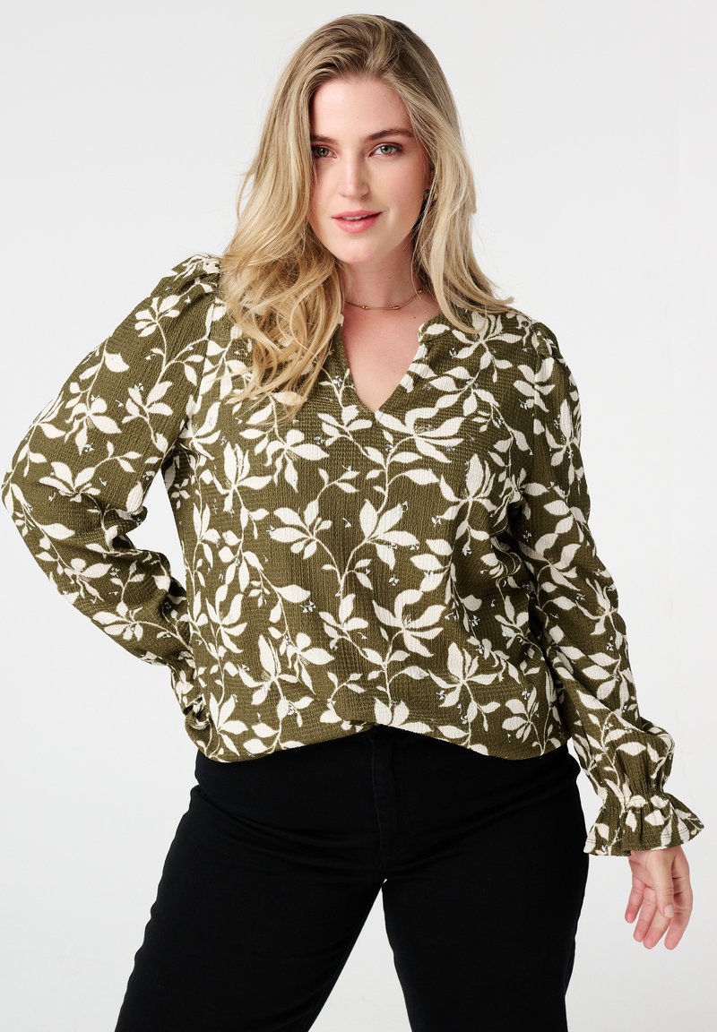 Blouse vert olive avec motif floral blanc, dotée d'un décolleté en V, tissu texturé et poignets froncés. Associée à un pantalon noir.
